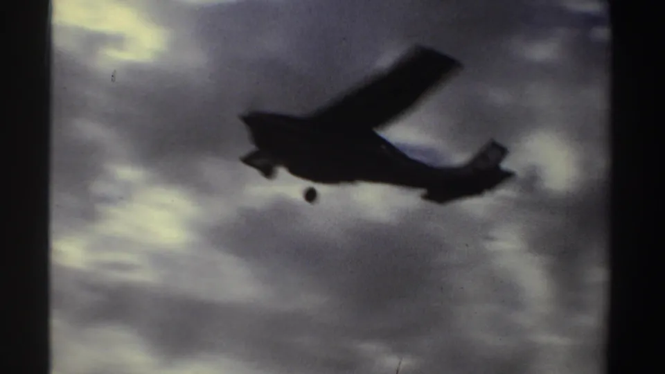 A small aircraft passes close overhead as a dark silhouette, its wing and landing gear visible against grey clouded sky.