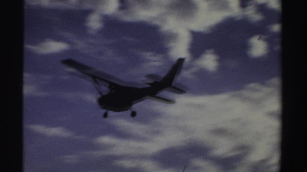 Small propeller aircraft with landing gear visible approaches across a cloudy sky in 1969 Sudan home-movie footage.