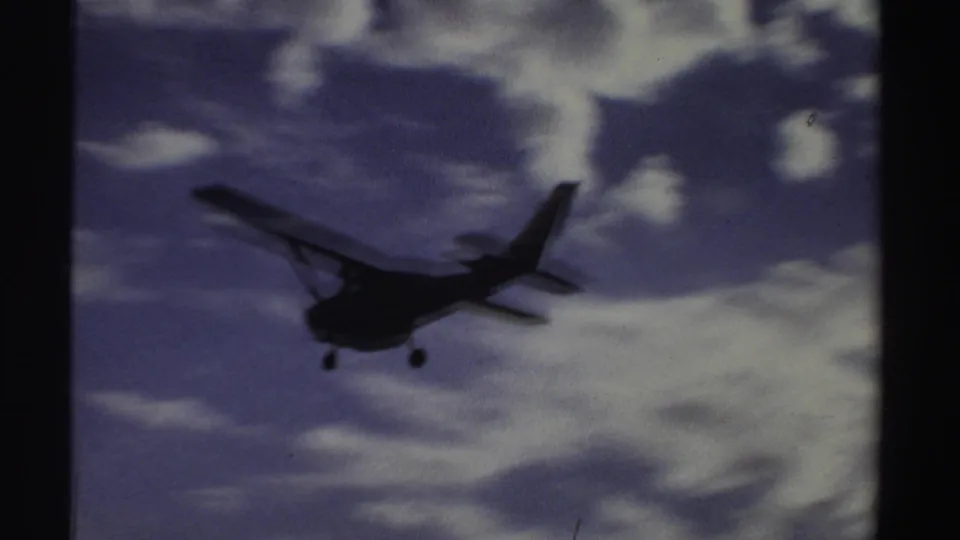 Small propeller aircraft with landing gear visible approaches across a cloudy sky in 1969 Sudan home-movie footage.