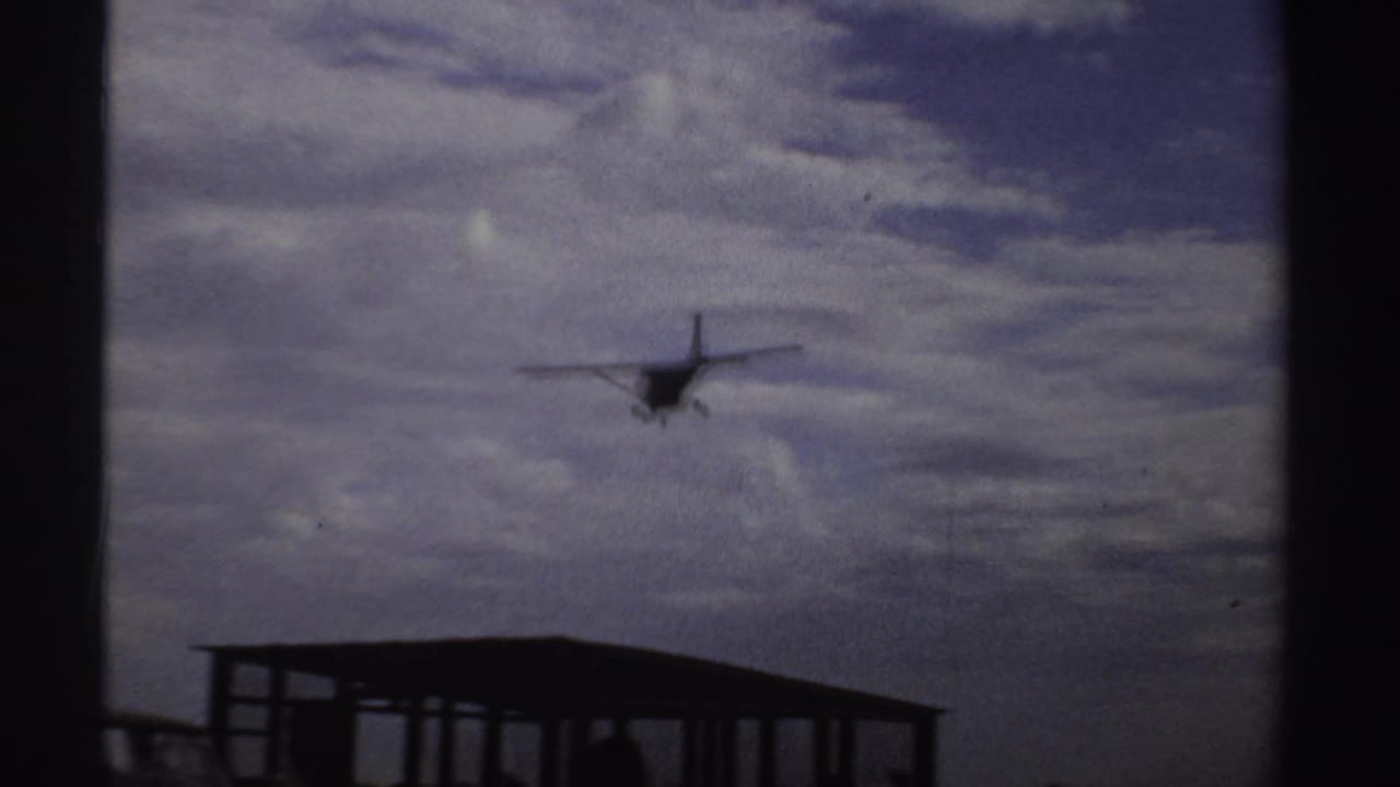Distant small aircraft descends over a simple open-sided shelter at a rural airstrip in 1969 Sudan footage.