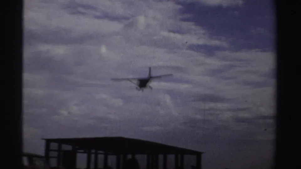 Distant small aircraft descends over a simple open-sided shelter at a rural airstrip in 1969 Sudan footage.