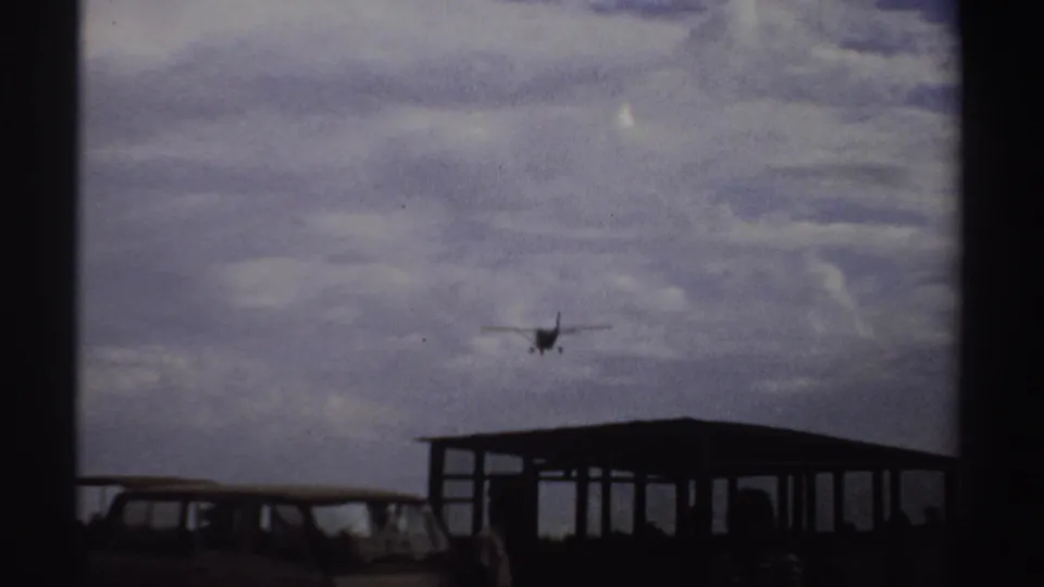 Small aircraft approaches over a parked vintage van and open-sided shelter, with distant silhouetted people near the airstrip.