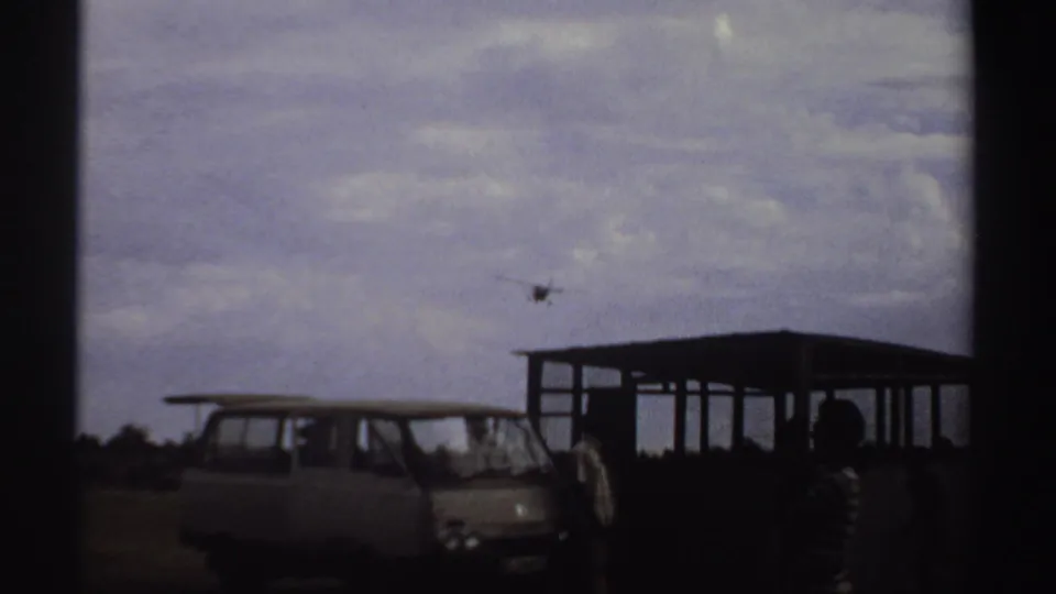 Small plane approaches low beyond a parked vintage van, open shelter, and silhouetted bystanders in 1969 Sudan airstrip footage.