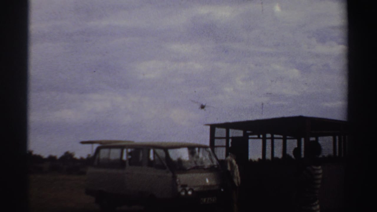 Small airplane above a parked van and open shelter at a rural airstrip.