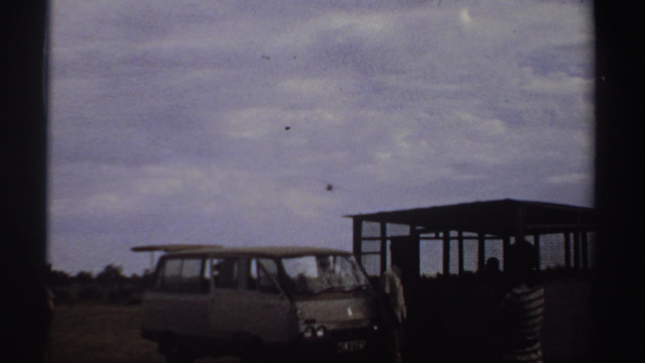 The aircraft crosses low over the rural Sudan airstrip scene, with a parked van and several onlookers beneath a cloudy sky.