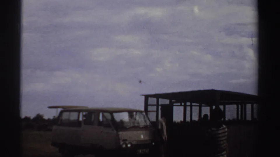 A small airplane recedes across the sky while the van, shelter, and spectators remain in the foreground of the Sudan airstrip footage.
