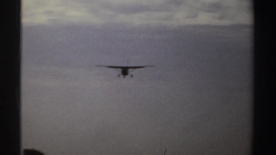 The approaching propeller aircraft is most visible near center frame, with open terrain below and cloudy sky above.