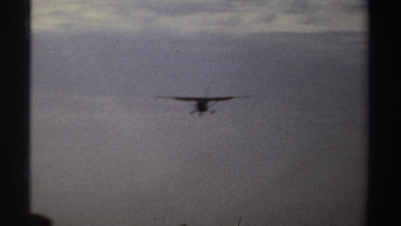1969 Sudan 8mm frame showing a small propeller airplane descending above a low treeline in soft, grainy light.