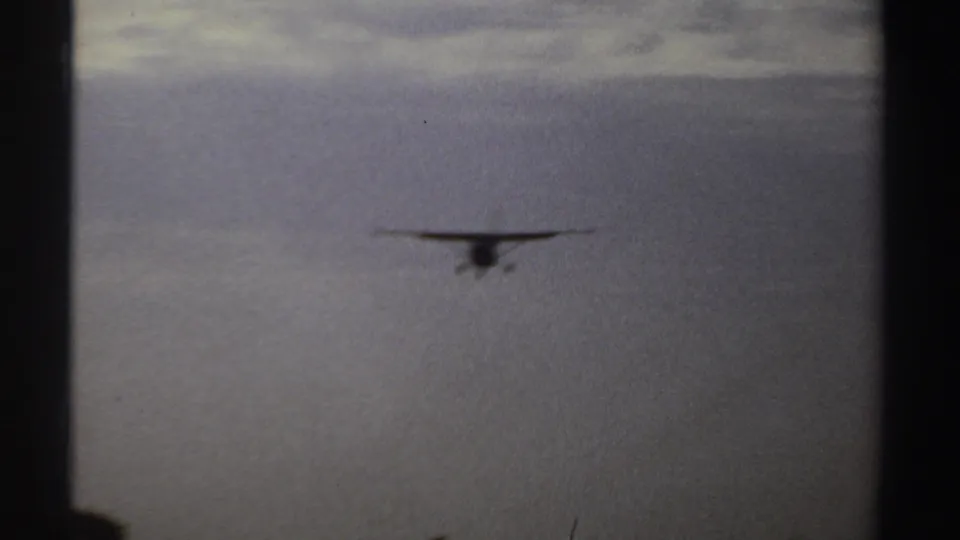 1969 Sudan 8mm frame showing a small propeller airplane descending above a low treeline in soft, grainy light.