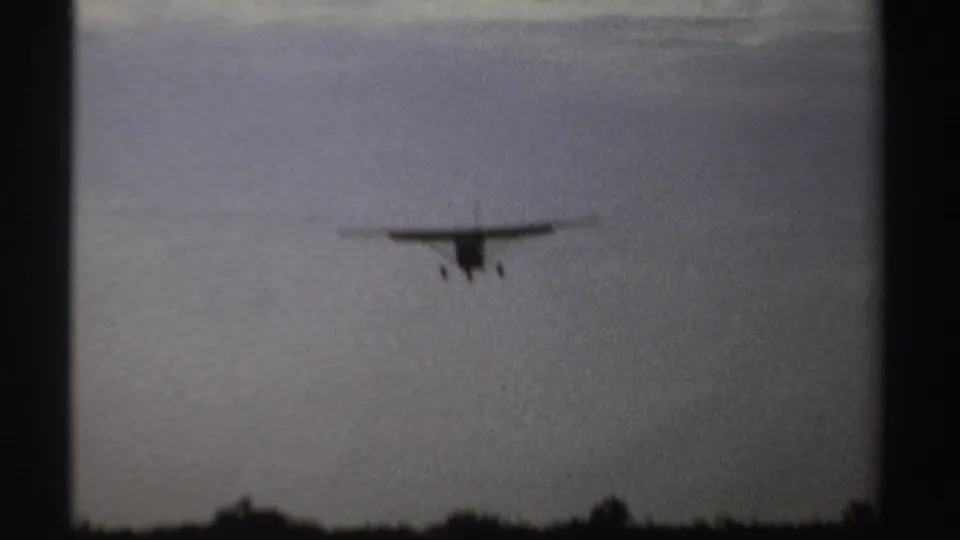 1969 Sudan 8mm frame with a small high-wing airplane on final approach over trees and open ground.