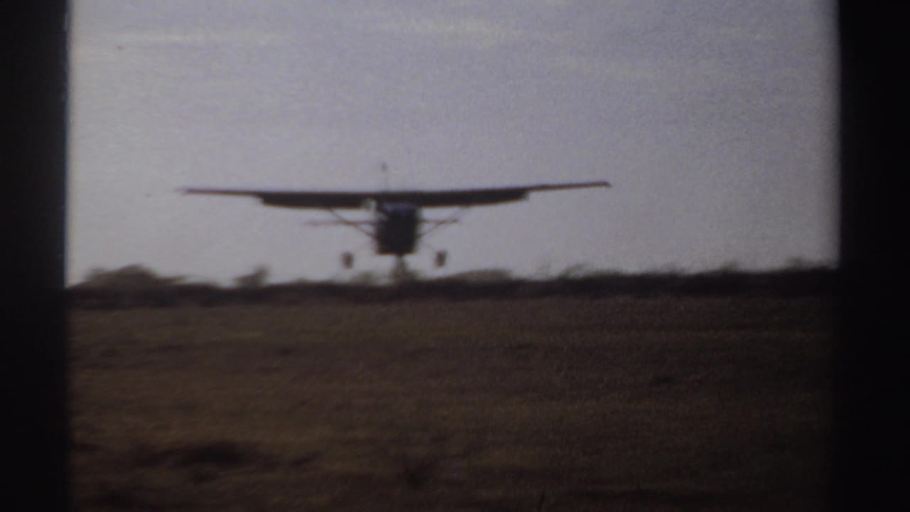 A small propeller aircraft approaches an open Sudan field in 1969, framed by dark 8mm gate edges.