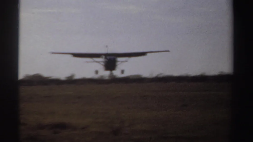 A small propeller aircraft approaches an open Sudan field in 1969, framed by dark 8mm gate edges.