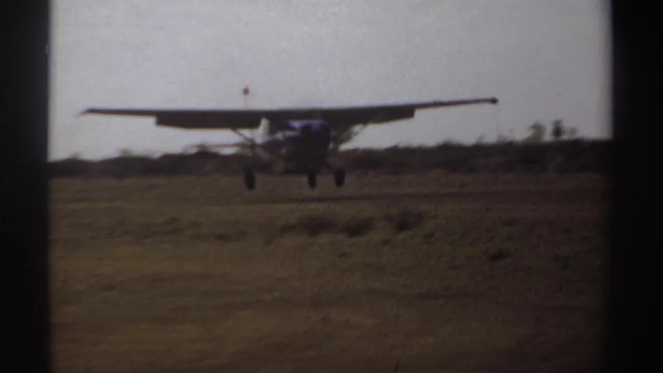 A blue and white high-wing aircraft descends toward a rough open landing field in Sudan, 1969.
