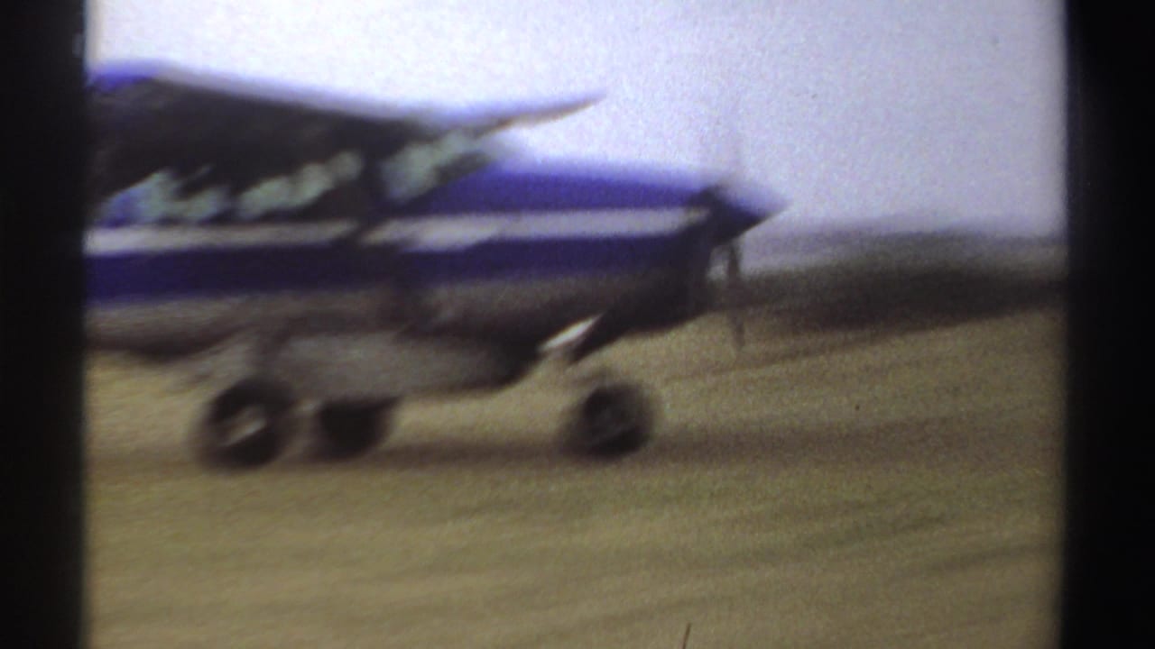 A blue and white single-engine plane taxis across a grassy Sudan airfield in 1969, seen in soft 8mm focus.
