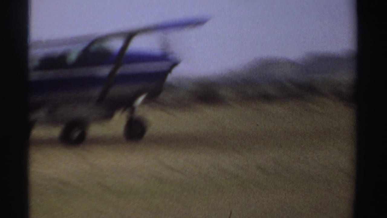Small blue aircraft seen side-on while taxiing across grass.