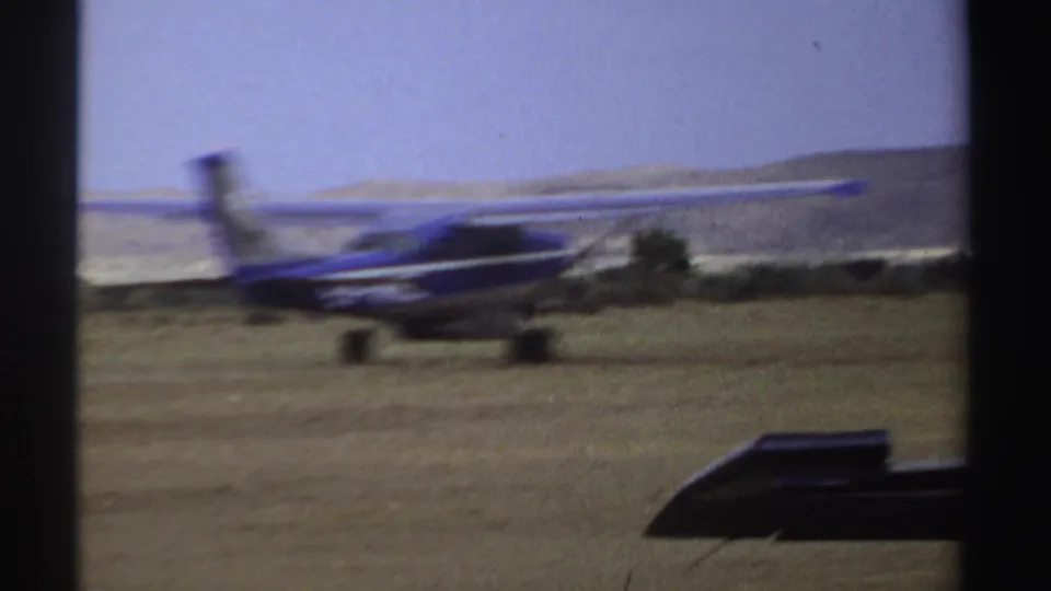 The blue plane continues across the grass field, framed against open sky, low distant hills, and a dark foreground edge.