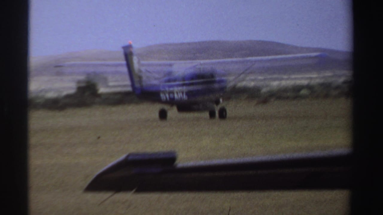 Small blue-and-white aircraft on a grass runway with hills and sky beyond.