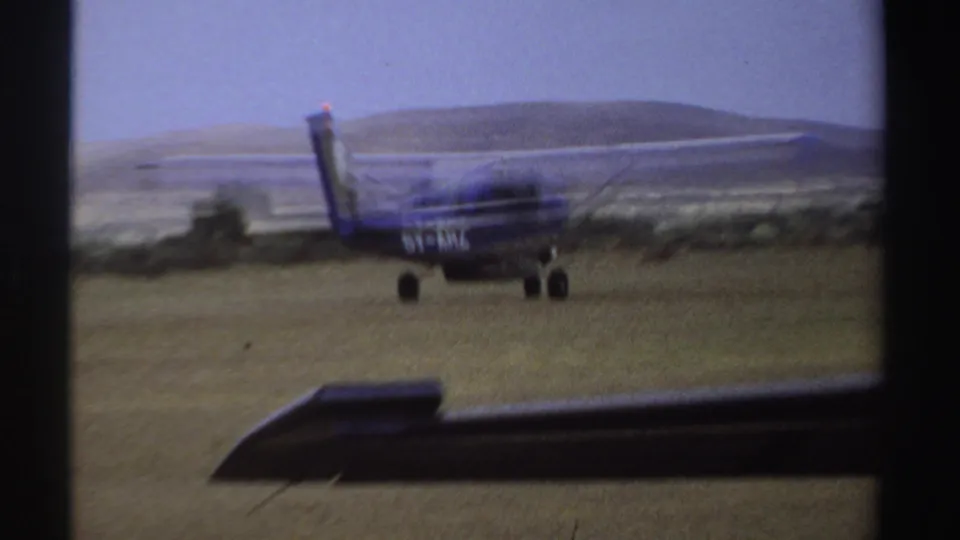 1969 Sudan 8mm frame showing a small blue-and-white aircraft near a grass runway, seen through dark foreground framing with distant hills.
