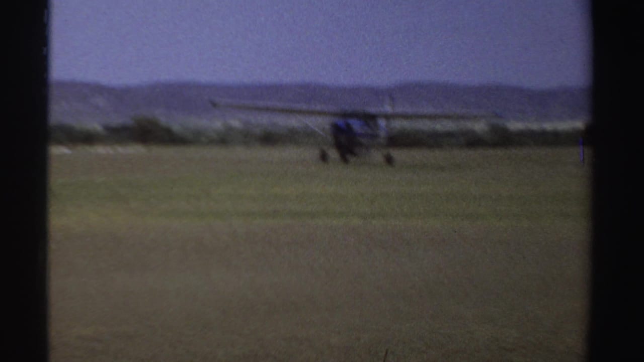 1969 Sudan 8mm frame of a small aircraft taxiing on a grass airstrip, with a windsock and low hills visible in the distance.