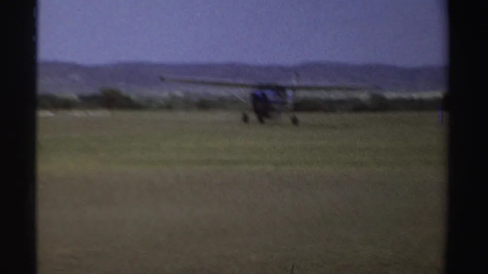1969 Sudan 8mm frame of a small aircraft taxiing on a grass airstrip, with a windsock and low hills visible in the distance.