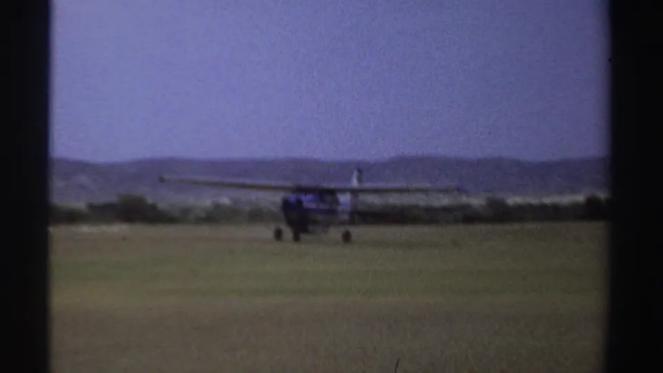 1969 Sudan 8mm frame showing the blue-and-white aircraft farther along the grass runway, with muted hills and sky beyond.
