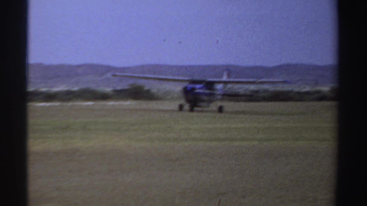 1969 Sudan 8mm frame showing a small blue and white airplane moving across a grass airstrip under a pale sky.