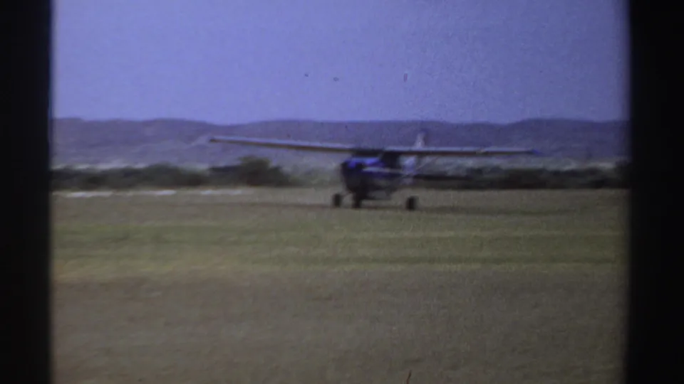 1969 Sudan 8mm frame showing a small blue and white airplane moving across a grass airstrip under a pale sky.