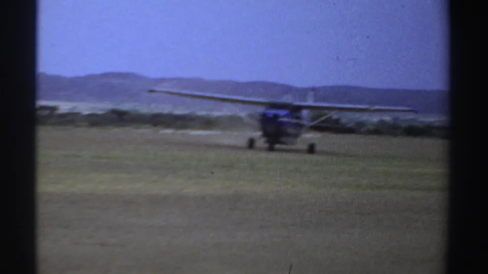 1969 Sudan 8mm frame with the small blue and white airplane near frame right on an open grassy airstrip.