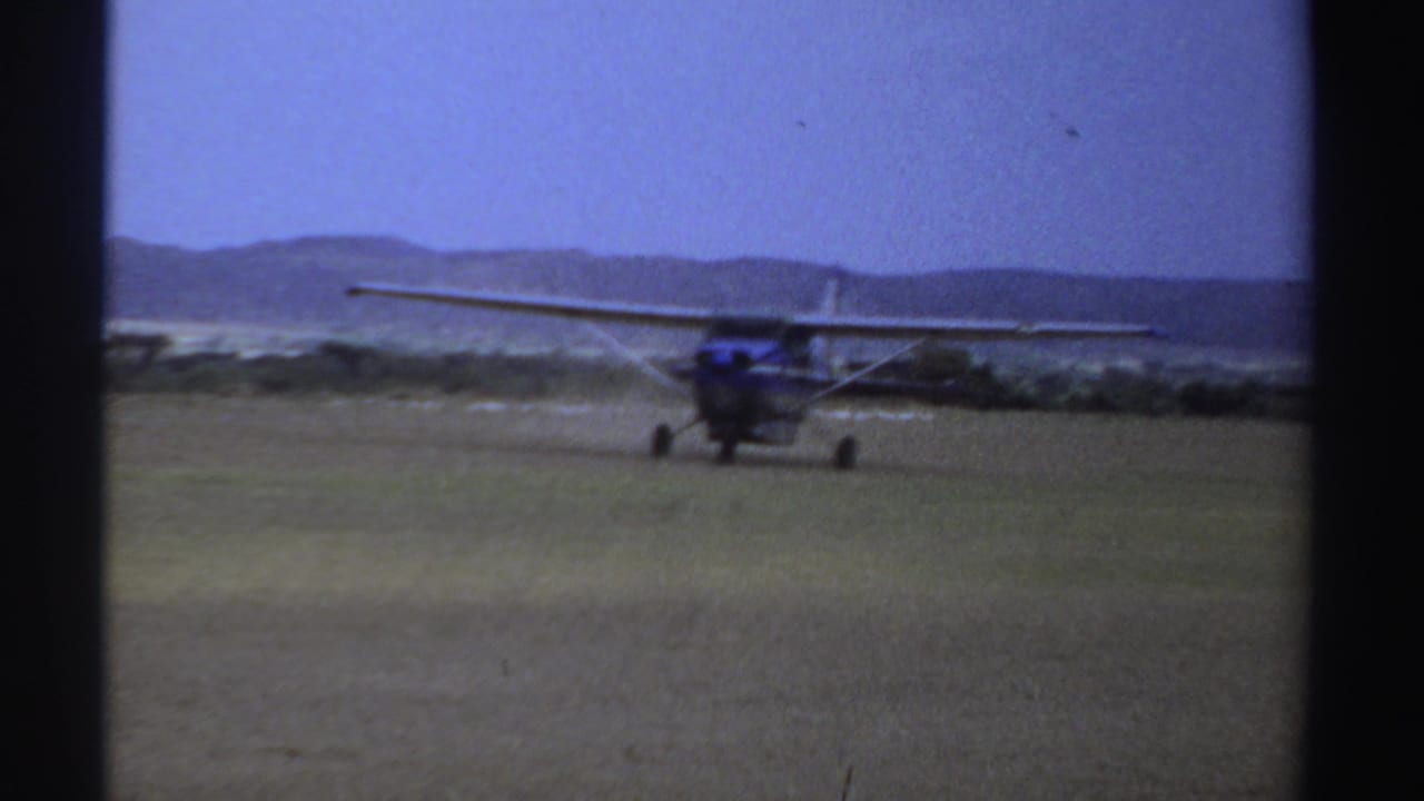 1969 Sudan 8mm frame showing a high-wing propeller plane moving across a grass runway under a pale blue sky.