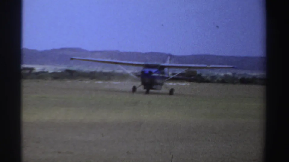1969 Sudan 8mm frame showing a high-wing propeller plane moving across a grass runway under a pale blue sky.