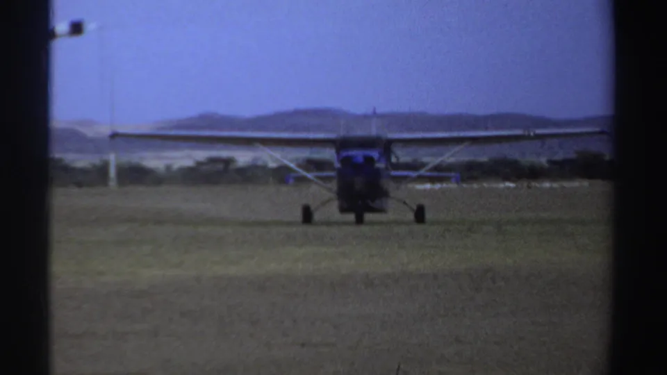 A centered 1969 Sudan home-movie frame shows the small high-wing plane taxiing away or toward camera on the grass strip.