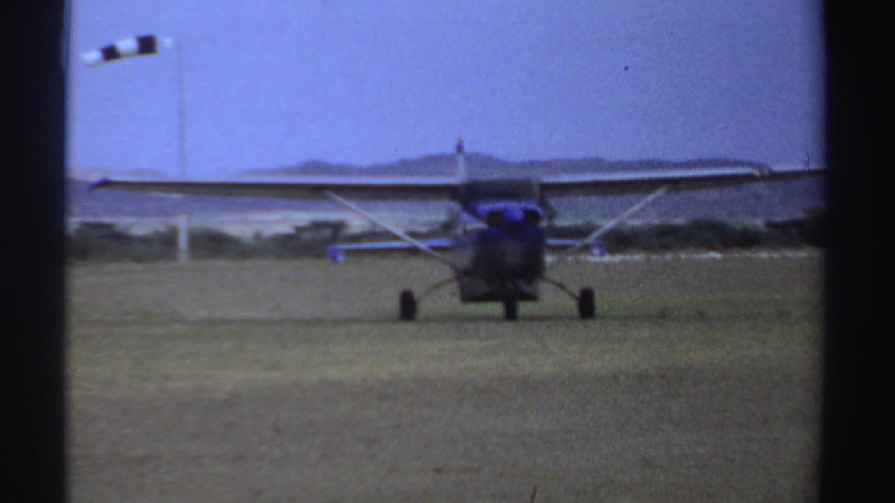 1969 Sudan home-movie frame of a small high-wing aircraft rolling on grass beneath a clear blue sky.