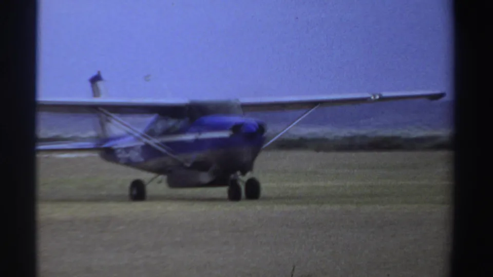 1969 Sudan frame with the small blue-and-white airplane seen from the side as it rolls across a grassy airfield.
