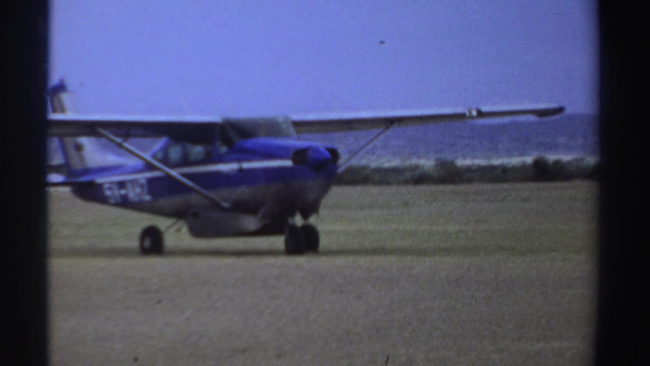 In Sudan archival 8mm footage from 1969, a small blue and silver high-wing plane taxis across a sparse dirt airfield.