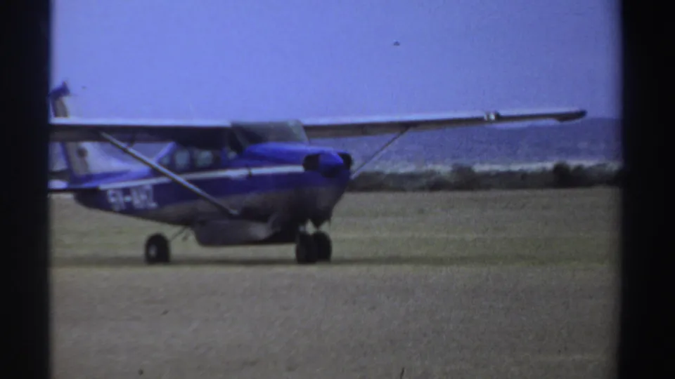 In Sudan archival 8mm footage from 1969, a small blue and silver high-wing plane taxis across a sparse dirt airfield.
