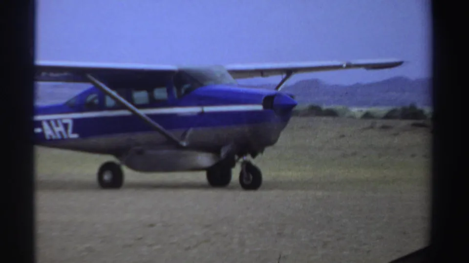 A closer side view shows the small blue aircraft continuing along the dirt runway in Sudan, with scrub and hills beyond.
