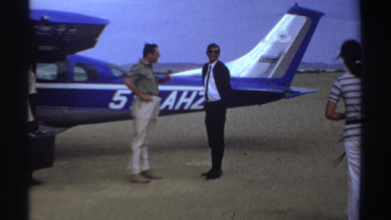 Two men stand beside a blue-and-white aircraft while a woman walks past in the foreground.