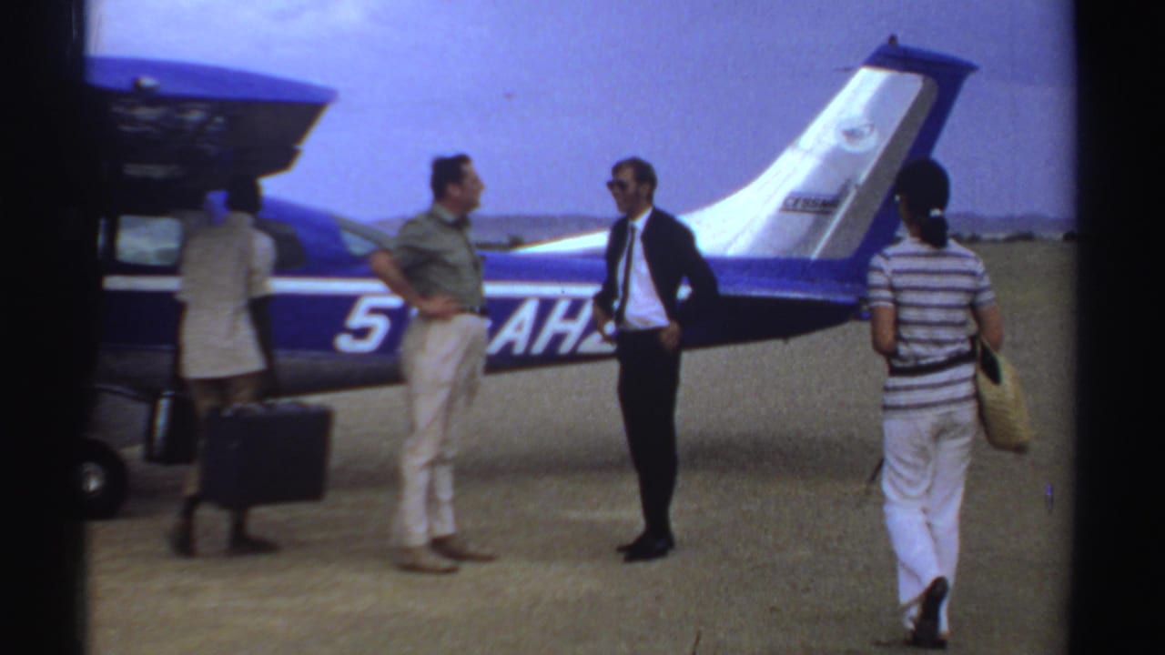 Travelers at a Sudan airfield stand by the open baggage area of a blue-and-white aircraft, with luggage and people moving around it.