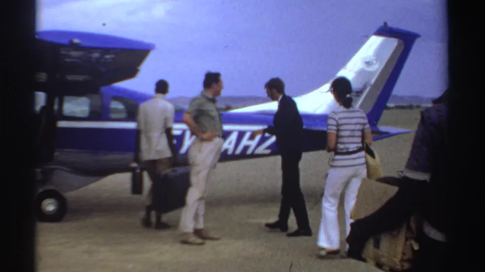 Several travelers in 1969 Sudan footage cluster around a single-engine aircraft, with one person handling luggage near the wheel.