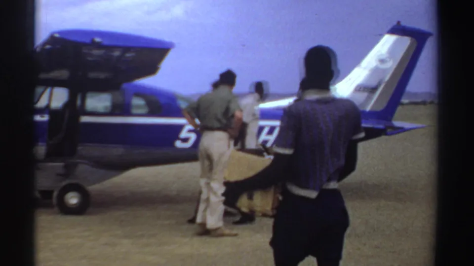 At a Sudan airfield, travelers walk beside the blue-and-white aircraft as the shot catches the tail, fuselage, and figures from behind.