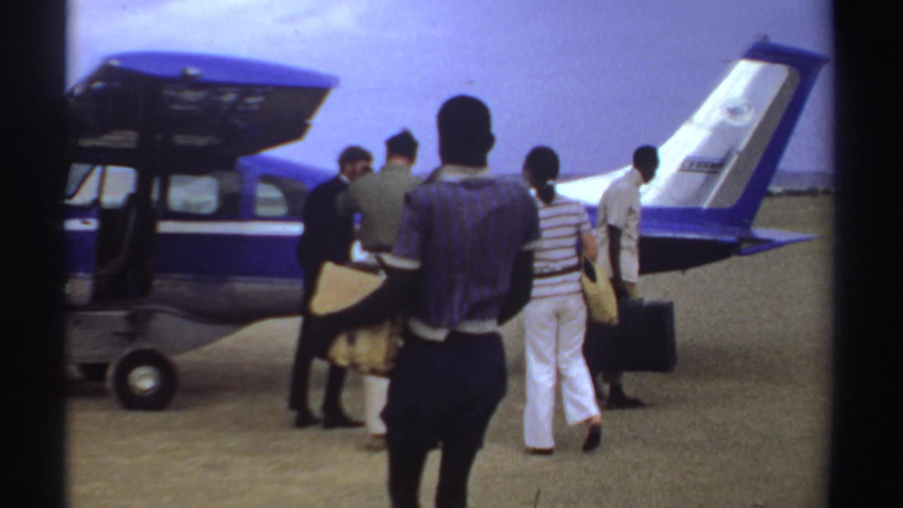 In Sudan 1969 home-movie footage, passengers with luggage approach a blue-and-white propeller plane on an open airfield.