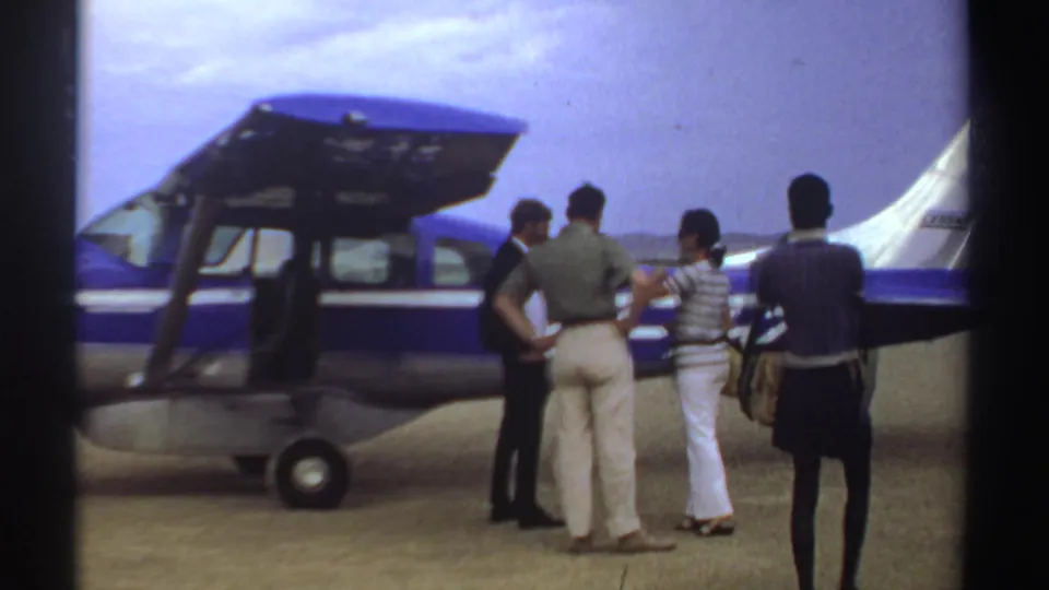 A Sudan 1969 airfield shot shows passengers beside the aircraft as a foreground figure watches with a bag over one shoulder.
