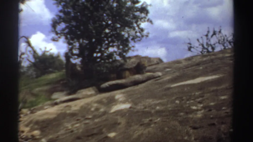1969 Sudan 8mm frame showing a tilted handheld view across rocky ground, bushes, a tree, and cloudy sky in open scrubland.