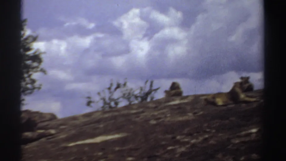 1969 Sudan 8mm frame showing cloudy sky over a dark rocky slope, sparse shrubs, and small rock forms on the ridge.