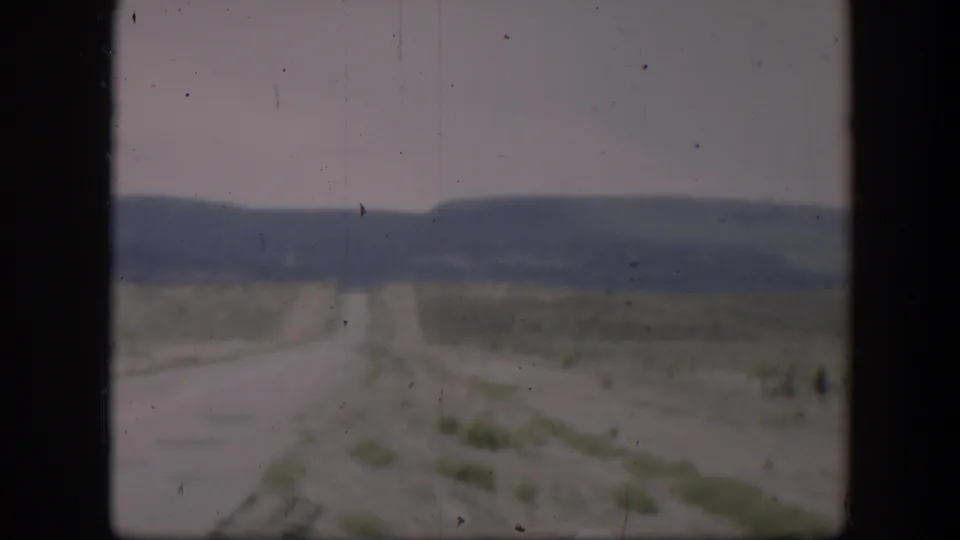 Long empty Utah desert road in 1951, bordered by sparse vegetation and framed by dark vehicle or film edges.