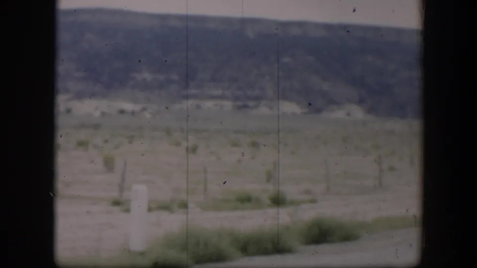 1951 Utah home-movie still showing roadside vegetation, a wire fence, open field, and distant low hills from a moving vehicle.