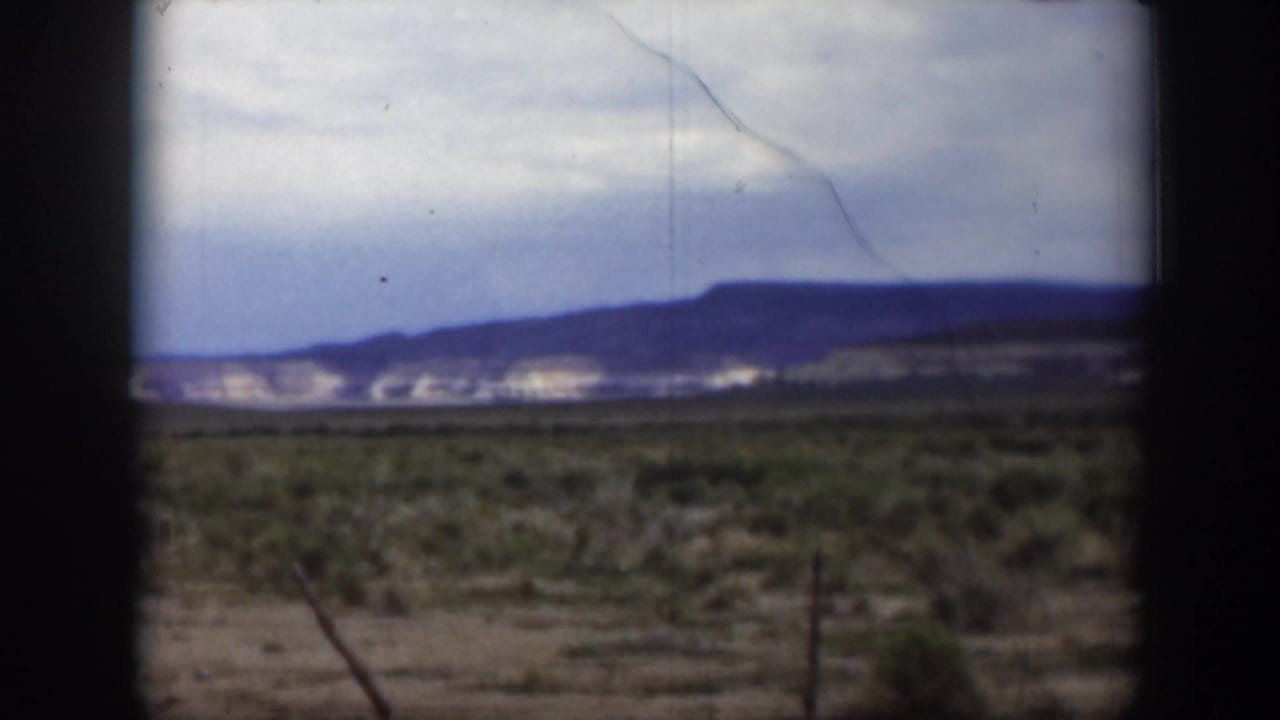 1951 Utah 8mm landscape frame with scrubby foreground, pale hillside patches, and a dark mountain ridge beneath an overcast sky.