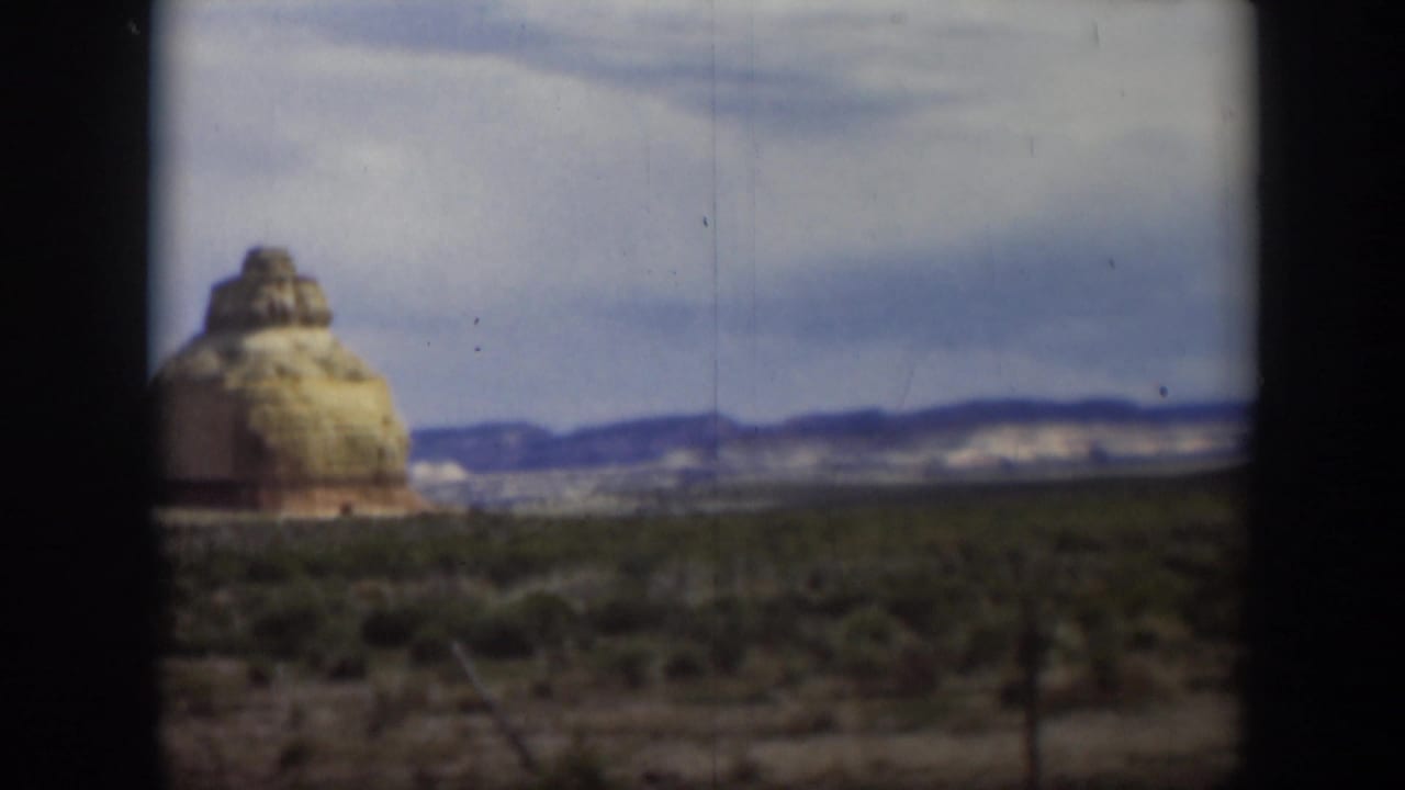 1951 Utah travel footage showing a small rock formation rising from desert scrub, with distant hills and clouded blue sky behind it.