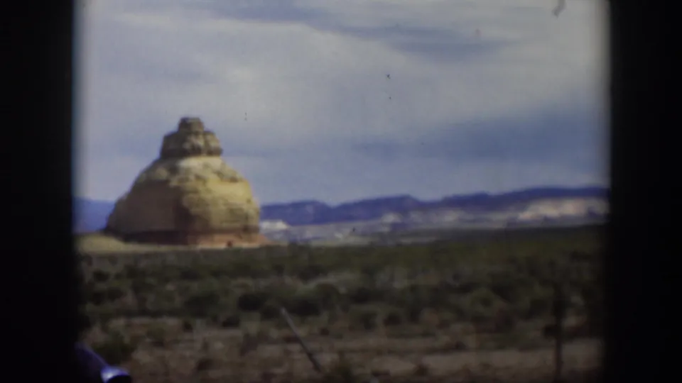 1951 Utah home-movie view through a vehicle opening toward a large tan rock formation with scrubland and distant hills.