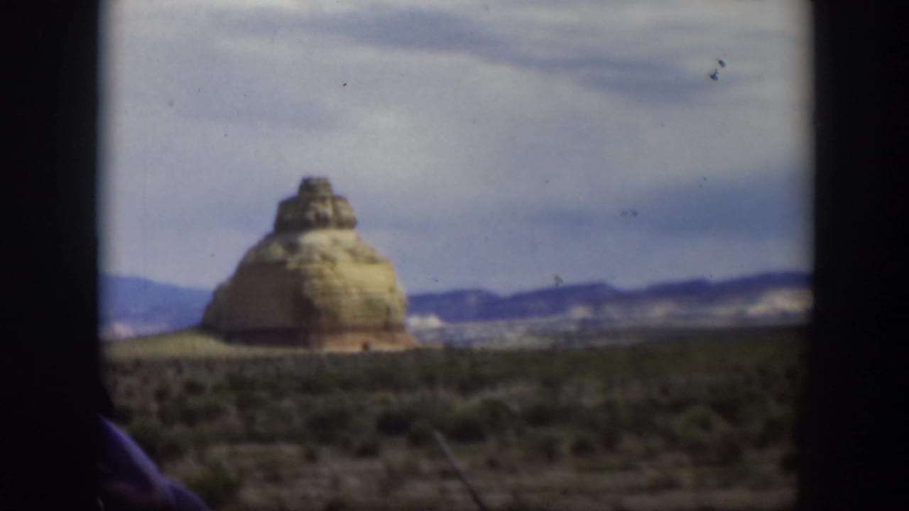 1951 Utah road-trip frame showing a tan desert rock formation, low vegetation, hills, and dark vehicle framing at the sides.